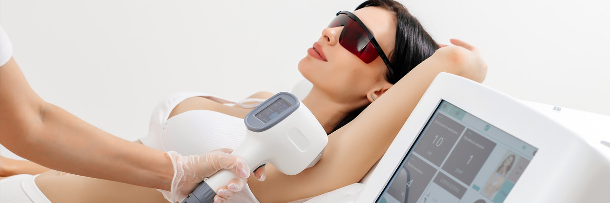 Body Care Concept. Laser Hair Removal. Young Caucasian Woman In Safety Glasses Lies On The Sofa, Female Cosmetologist Doing Laser Hair Removal In A Beauty Salon
